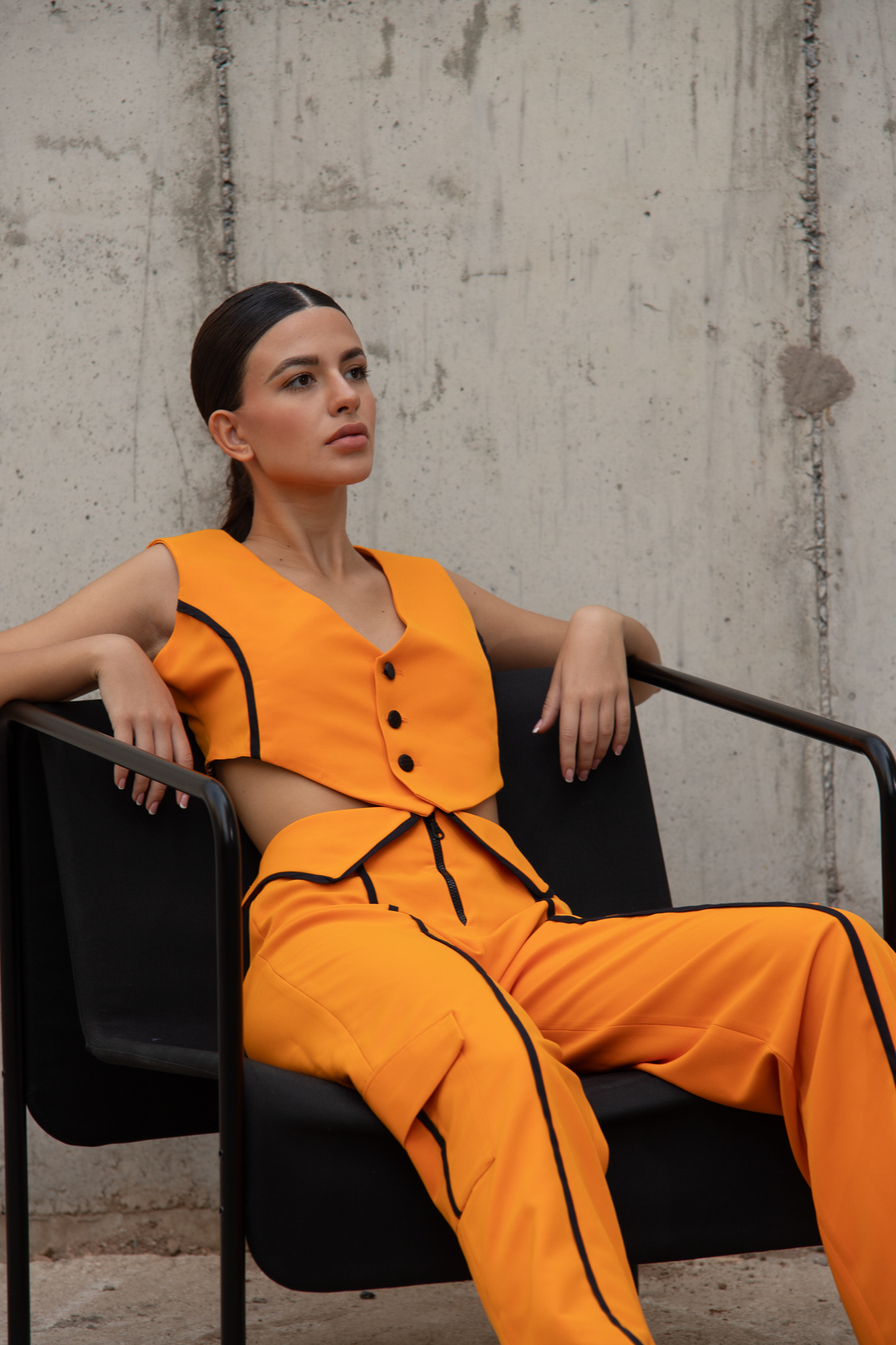 A brunette girl sitting on a chair, with an orange vest, with black details. Lace up back and buttons on the front side and orange , straight line trousers with collar on the waist, zipper and black-stripe details.