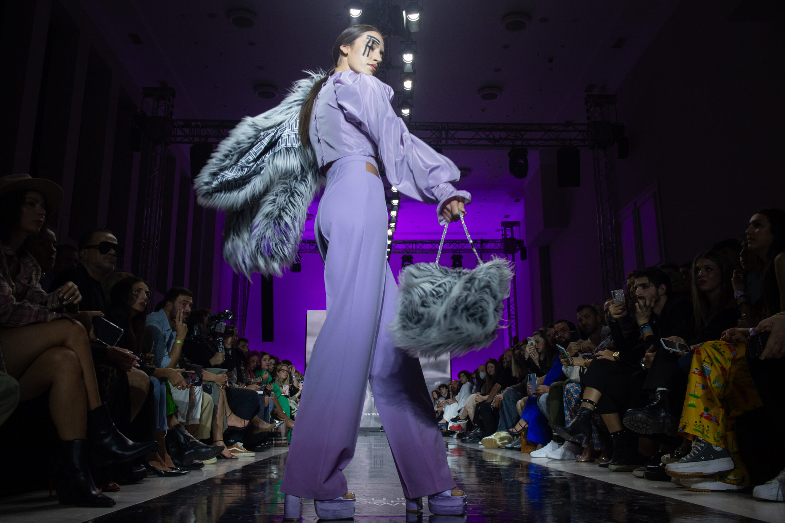 A model posing on catwalk with lilac set, a cropped top with long puffy sleeves holding an faux fur bag and a faux fur coat at Athens Fashion Week.